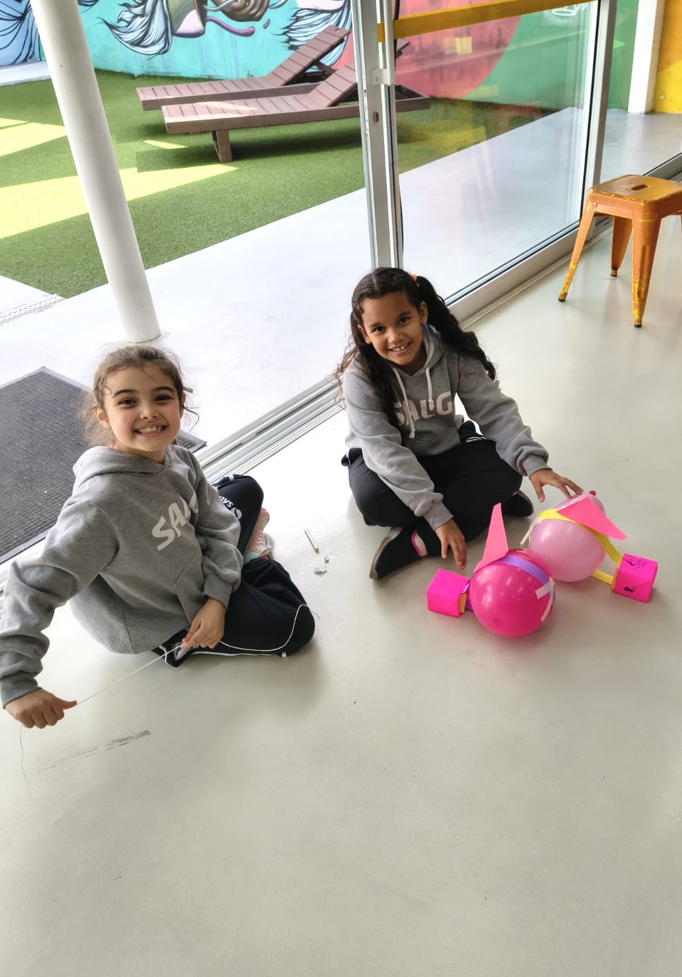 Duas meninas sorrindo ao lado de seus foguetes de bexiga rosa na atividade de Cultura e Inovação
